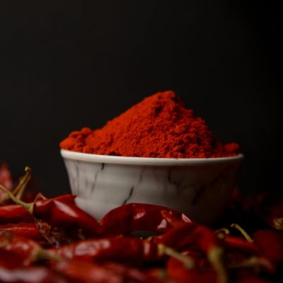 A bowl of vibrant red chili powder surrounded by whole dried chilies on a dark background.