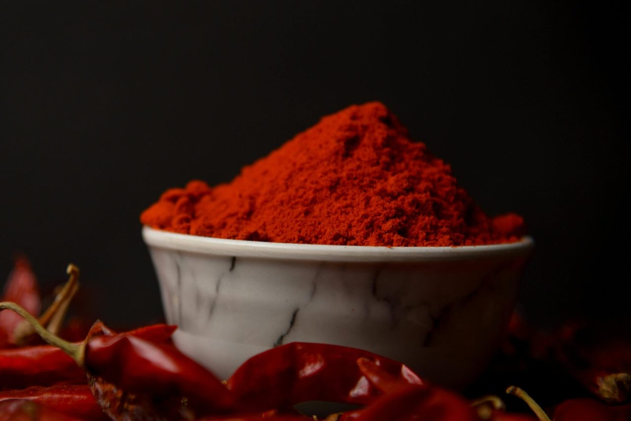 A bowl of vibrant red chili powder surrounded by whole dried chilies on a dark background.