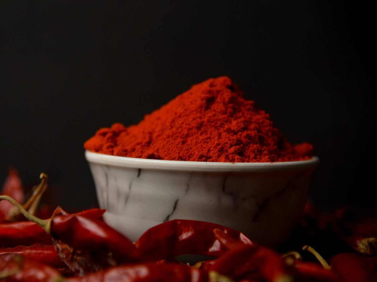 A bowl of vibrant red chili powder surrounded by whole dried chilies on a dark background.